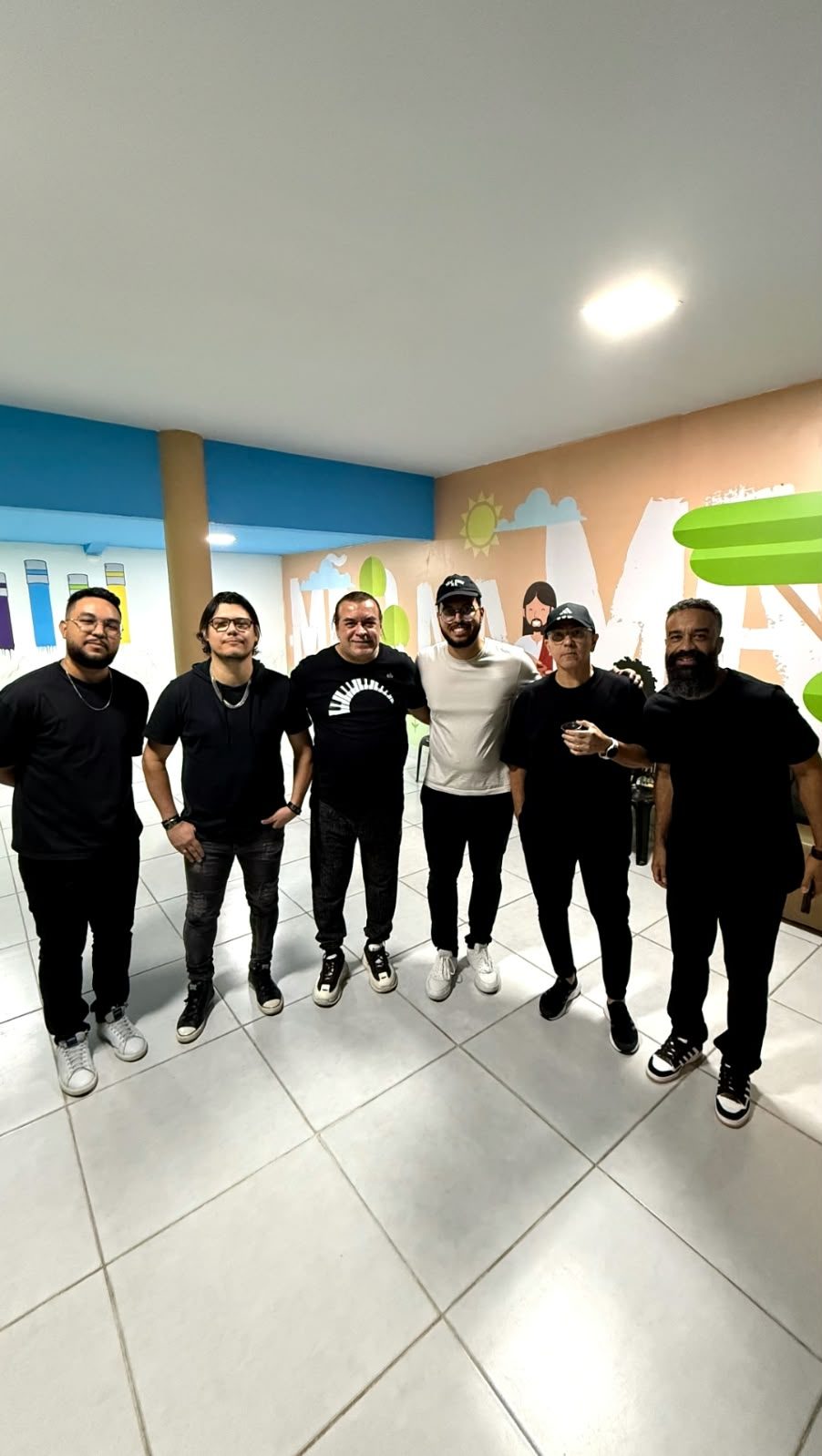 Seis homens de preto lado a lado num salão com mural